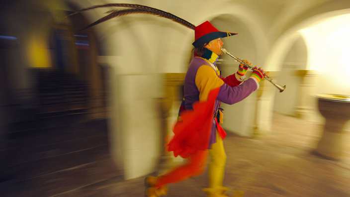 Ein Mann in bunter, mittelalterlicher Kleidung. Er trägt einen Federhut. Er hält ein Blasinstrument in der Hand und wandert durch alte Gewölbe.