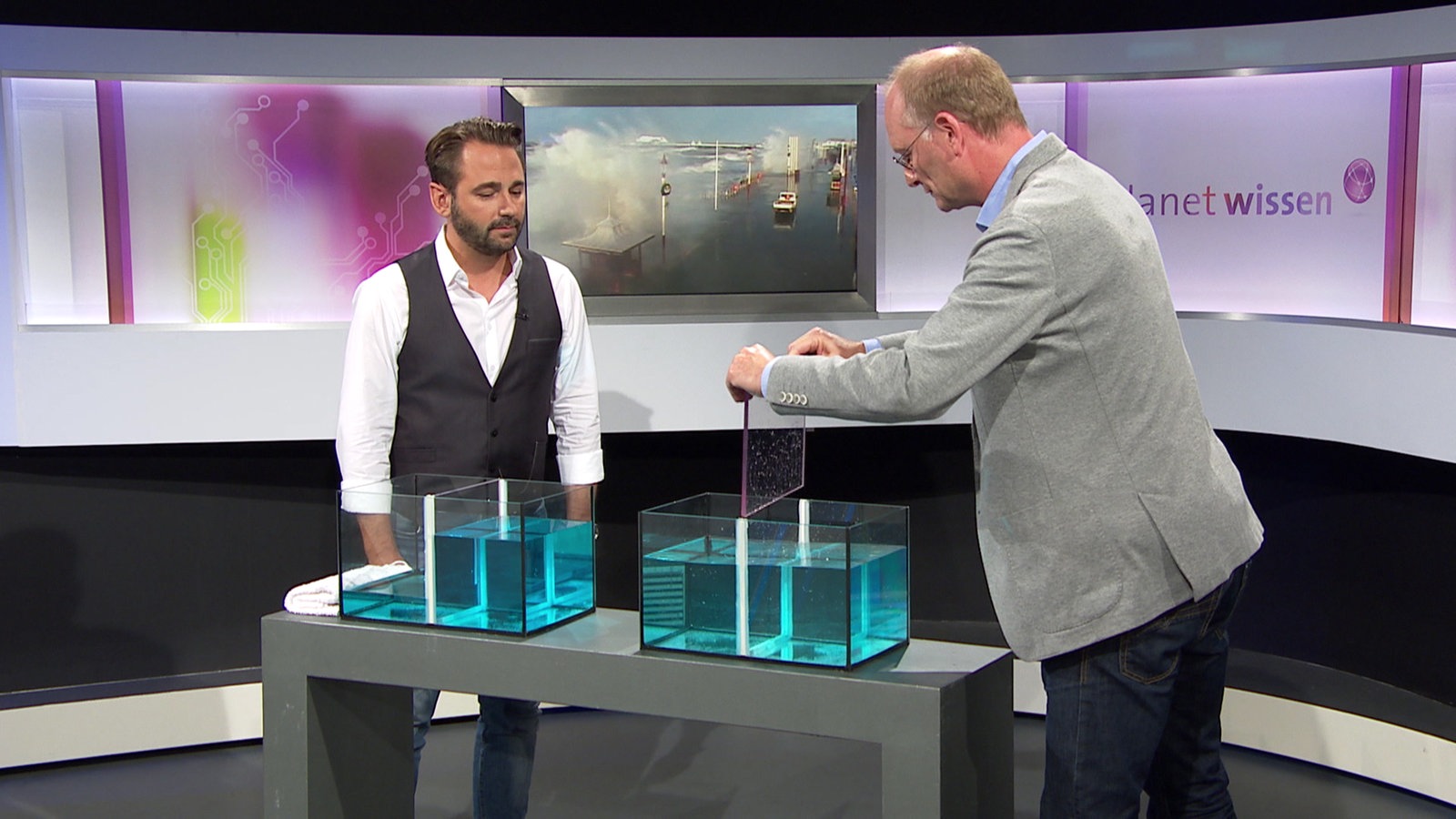 Sven Plöger und Dennis Wilms stehen im Studio an einem Tisch mit zwei wassergefüllten Plexiglaskästen.