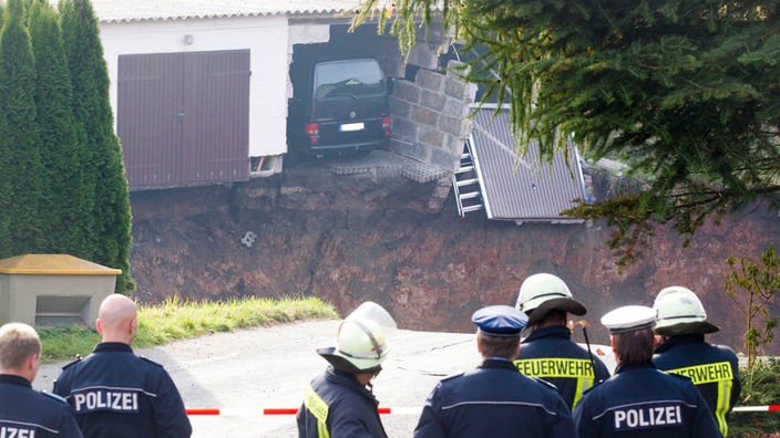 Loch vor Garage mit Auto, davor Polizisten an Absperrung