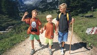 Drei Jungs gehen einen Wanderweg entlang, hinter ihnen erstrecken sich die Alpen.