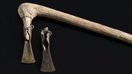Zwei bronzene Beilklingen, eine mit Holzhalterung vor schwarzem Hintergrund.