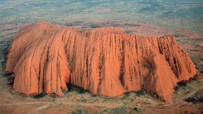Blick von oben auf den aus rotem Sandstein bizarr geformten Ayers Rock, der inmitten einer flachen, kargen Steppenlandschaft liegt.