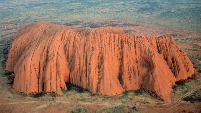 Blick von oben auf den aus rotem Sandstein bizarr geformten Ayers Rock, der inmitten einer flachen, kargen Steppenlandschaft liegt. | Bildquelle: dpa Picture-Alliance/Lehtikuva Blick von oben auf den aus rotem Sandstein bizarr geformten Ayers Rock, der inmitten einer flachen, kargen Steppenlandschaft liegt.
