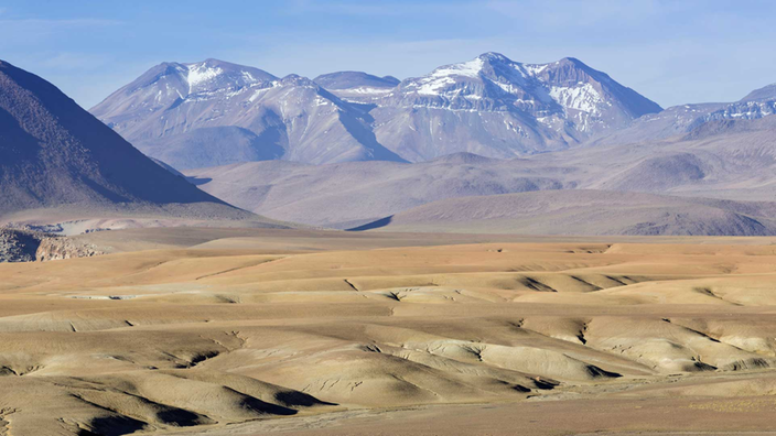 Hochland der Atacamawüste bei San Pedro de Atacama, Chile. Es sind sanfte, trockene Gesteinsfaltungen zusehen. Im Hintergrund ragen mit Schnee bedeckte Gipfel herauf.
