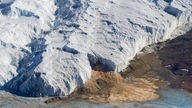 Links ist eine große Eisschicht in der Antarktis von oben zu sehen, rechts davon liegt ein Trockental mit kargem, braunem Boden.