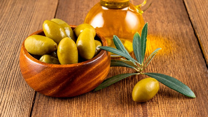 Grüne Oliven mit Olivenzweig in einer Holzschale und eine Flasche Olivenöl auf einem rustikalen Holzhintergrund.