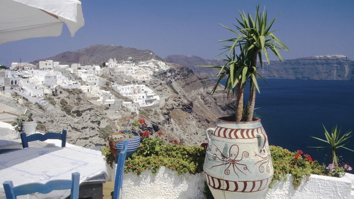 Blick über eine Terrasse auf weiße Häuser der Insel Santorin, die an einem Kraterrand zu kleben scheinen.