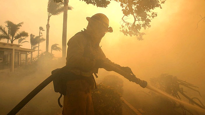 Ein Feuerwehrmann steht in dichtem Rauch und bekämpft Flammen mit einem Wasserschlauch. Die Luft schimmert rötlich vom Feuerschein.