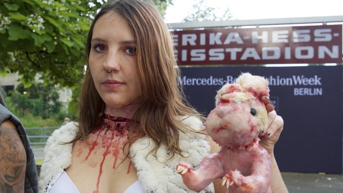 Das Bild zeigt eine Frau mit einer Kunstblutwunde am Hals. In der linken Hand hält sie eine Nachbildung eines Kaninchens, dessen Fell abgezogen wurde.