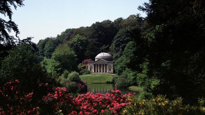 Ein dicht bewachsener Park mit See. Im Hintergrund steht ein kleiner griechischer Tempel.