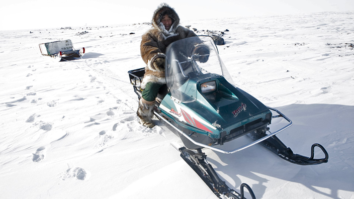 Ein Inuit in Fellkleidung auf einem Motorschlitten mit dem er einen zweiten Schlitten zieht unterwegs im endlosen Schnee und Eis der Arktis.