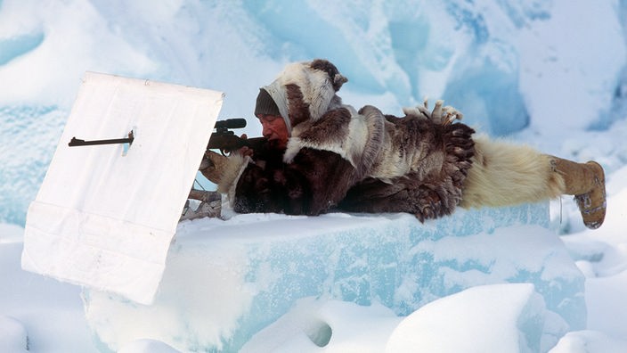 Ein warm in Pelz eingepackter Eskimo auf der Jagd; er liegt dabei mit dem Gewehr im Anschlag auf einem großen bläulichen Eisquader.