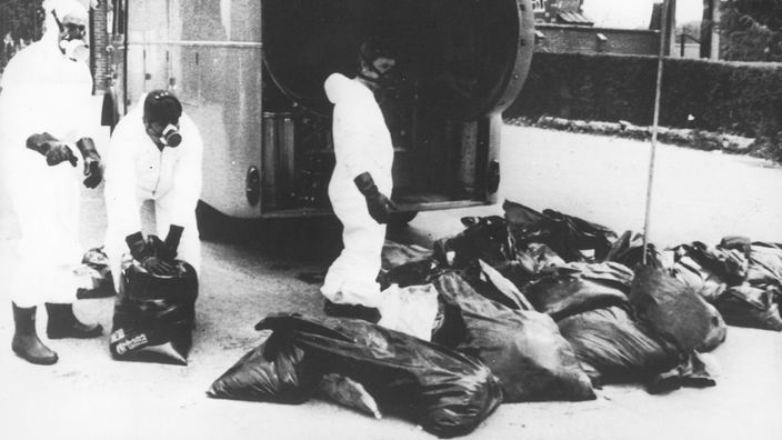 Schwarzweiß-Bild: Drei Männer in weißen Schutzanzügen mit Müllsäcken