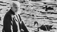 Konrad Lorenz sitzt am Wasserrand und dreht sich in Richtung Kamera, im Hintergrund schwimmt eine Ente und ein Schwan