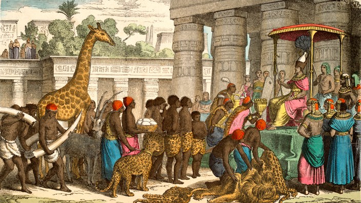 Kolorierter Holzstich:  Eine dunkelhäutige Menschenmenge bringt dem Pharao eine Giraffe, Raubtiere und Elefantenstoßzähne als Tribut