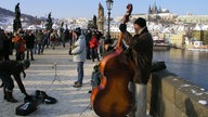 Zu sehen ist eine historische Brücke, die mit mehreren Stauten verziert ist. Auf ihr spazieren viele Menschen; im Vordergrund steht ein Straßenmusiker mit einem Kontrabass.