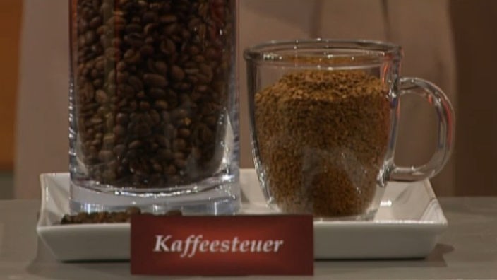 Glas mit Kaffeebohnen und löslichem Kaffee. Davor steht eine Schild auf "Kaffeesteuer" steht.