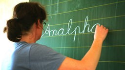 Eine Frau steht an der Tafel und schreibt mit Kreide das Wort "Analphabetismus" an 