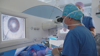 Ein Arzt, in blau gekleidet, lasert die Augen einer Patientin. Er schaut durch ein Vergrößerungsglas. Links auf einem Bildschirm wird die Pupille der Patientin vergrößert.