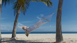 Eine Frau liegt am Strand unter Palmen in der Hängematte. 