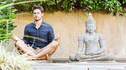 Ein Mann sitzt im Schneidersitz neben einer Buddhastatue und meditiert