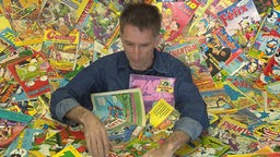 Jürgen Meyer, ein Mann mit aschigem Haar und in Jeanshemd, steckt bis zum Bauch in seiner Comicsammlung.