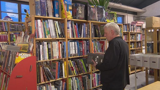 Ein Mann mit hellen Haaren und schwarzem Pullover steht in einem Comic-Geschäft und blättert in einem Heft.
