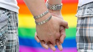 Zwei schwule Männer halten an den Händen. Im Hintergrund eine Regenbogenflagge. 