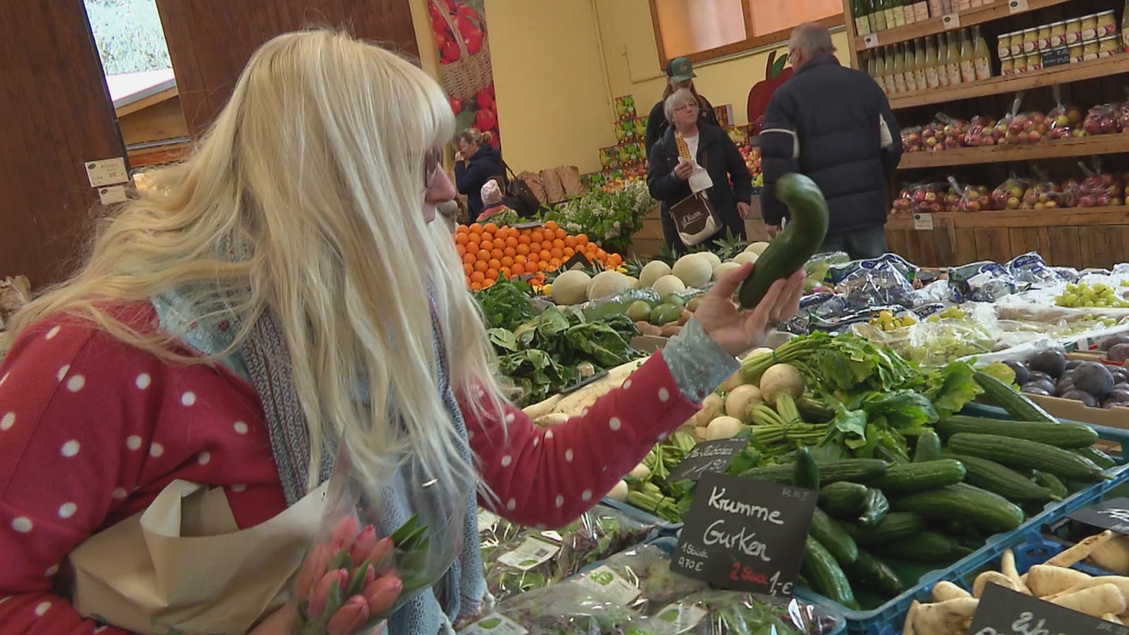 Eine Frau mit langen blonden Haaren steht in einem Geschäft und hat den Blick auf das Gemüsesortiment gerichtet. Sie hält eine krumme Gurke in der linken Hand.