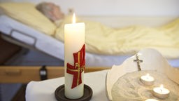 brennende Kerzen stehen auf einem Tisch, im Hintergrund liegt ein älterer Mann in einem Bett.