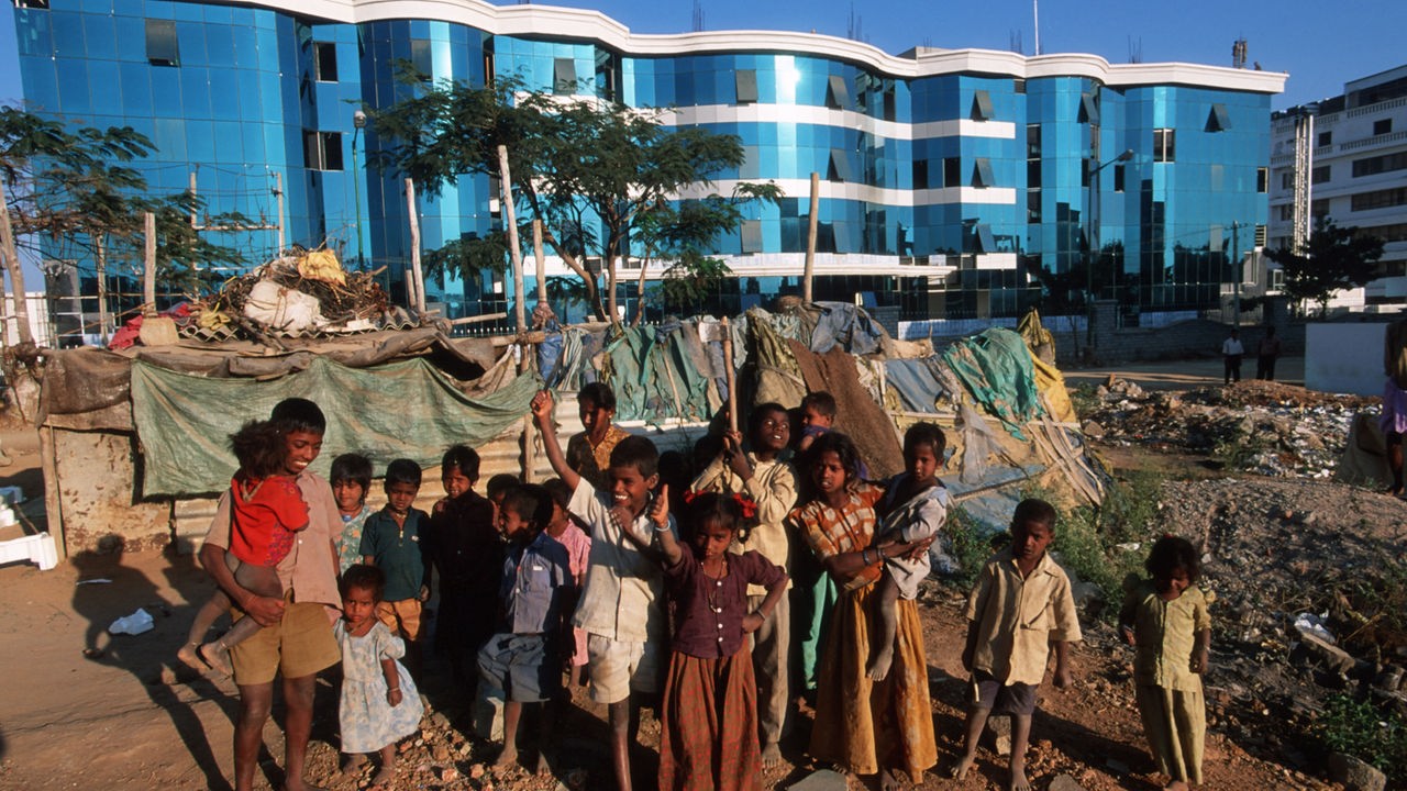 Eine Gruppe indischer Kinder steht barfuß in einem Zeltlager. Dahinter sieht man die moderne Architektur Bangalores. 