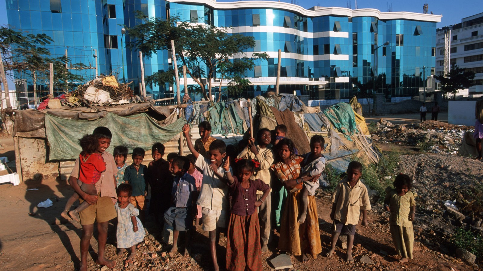 Eine Gruppe indischer Kinder steht barfuß in einem Zeltlager. Dahinter sieht man die moderne Architektur Bangalores. 