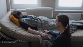 Eine ältere Frau liegt auf einem Bett mit Kopfhörer und Schlafmaske. Neben ihr sitzt eine junge Frau, die sie anschaut.