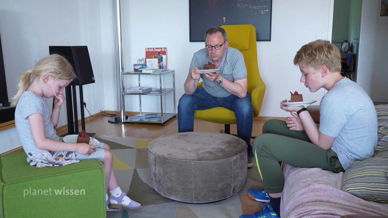 Ein Vater sitzt mit seiner Tochter und seinem Sohn kuchenessend um einen kleinen Plüschtisch.