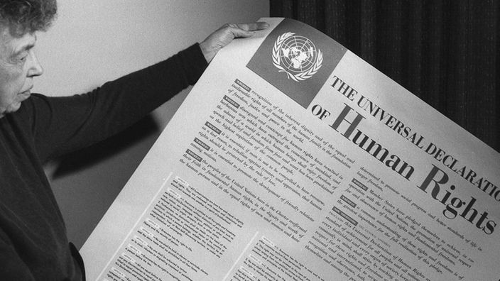 schwarz-weiß Aufnahme einer älteren Frau, die eine Zeitung mit der Überschrift "Human Rights" in der Hand hält.
