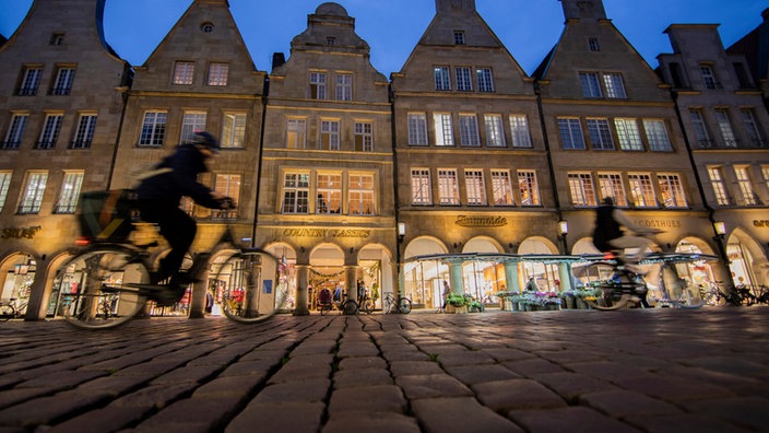 Zwei Fahrradfahrer vor der abendlich beleuchteten Kulisse des Prinzipalmarkts in Münster | Bildquelle: picture alliance / dpa Zwei Fahrradfahrer vor der abendlich beleuchteten Kulisse des Prinzipalmarkts in Münster