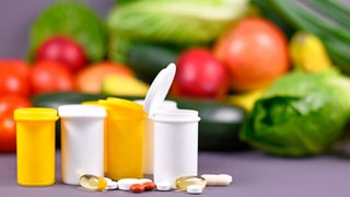 Döschen mit offenem Deckel vor Gemüse, daneben einzelne Nahrungsergänzungs-Pillen 