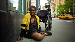 Eine Afroamerikanerin sitzt auf der Straße, im Hintergrund ein Rollstuhl und die Skyline New Yorks