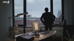 Ein Mann im Gegenlicht vor einem Fenster, im Vordergrund ein Schreibtisch mit Laptop