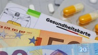 verschiedene Tabletten liegen neben einer Gesundheitskarte und zwei Geldscheinen.