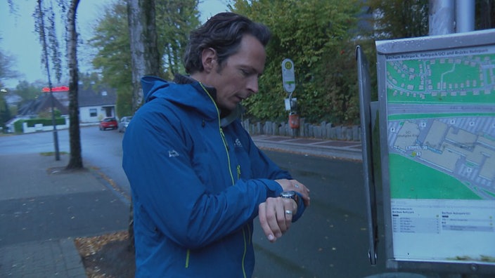 Moderator Jo Hiller blickt an einer Bushaltestelle auf die Uhr