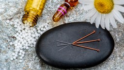Ein Fläschchen mit Globuli, eine Ampulle, eine Kamillenblüte und Akupunkturnadeln nebeneinander