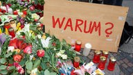 Schild mit Aufschrift "WARUM?" mit Kerzen und Blumensträußen.