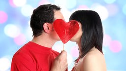 Symbolbild: Ein Liebespaar küsst sich. Der Mann hält einen überdimensionalen Lutscher in Herzform vor die Gesichter