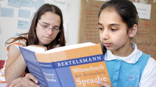 Zwei junge Mädchen sitzen in einem Klassenzimmer an einem Tisch, eine hält ein Deutsch-Wörterbuch in der Hand und liest darin.