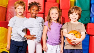 Vier Kinder stehen mit einem Ball vor bunten Matten in einer Turnhalle