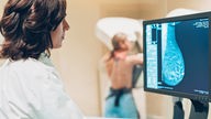 Eine Ärztin schaut auf das Bild einer Mammographie auf ihrem Monitor, im Hintergrund steht die Patientin am Untersuchungsgerät.