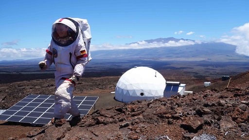 Frau im Raumanzug. Im Hintergrund ein weißer Kuppelbau in der Vulkanlandschaft Hawaiis 
