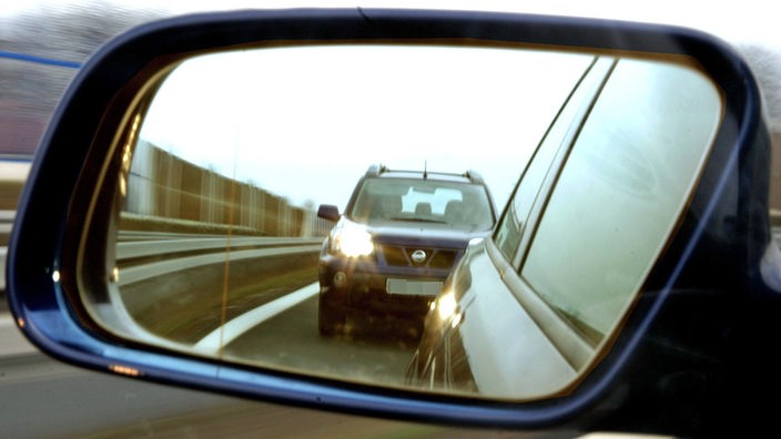 In dem Außenspiegel eines Kraftfahrzeugs ist ein zu dicht auffahrendes Auto mit Lichthupe zu sehen