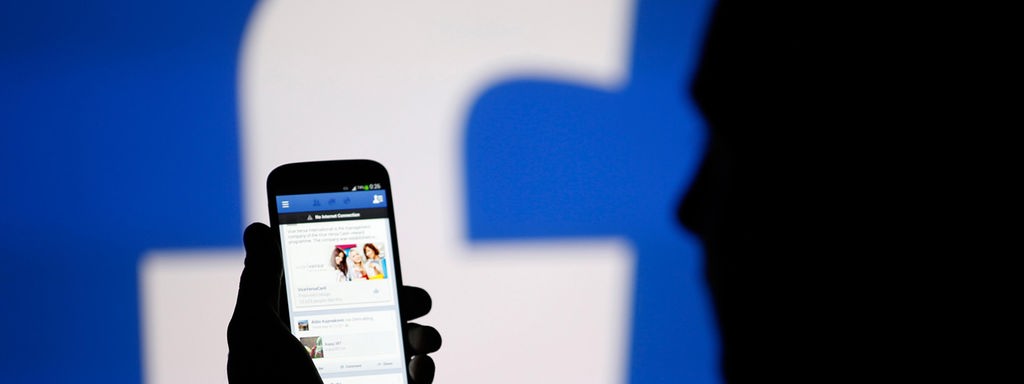 Silhouette eines Mannes, der vor einem großen Facebook-Logo auf sein Smartphone schaut